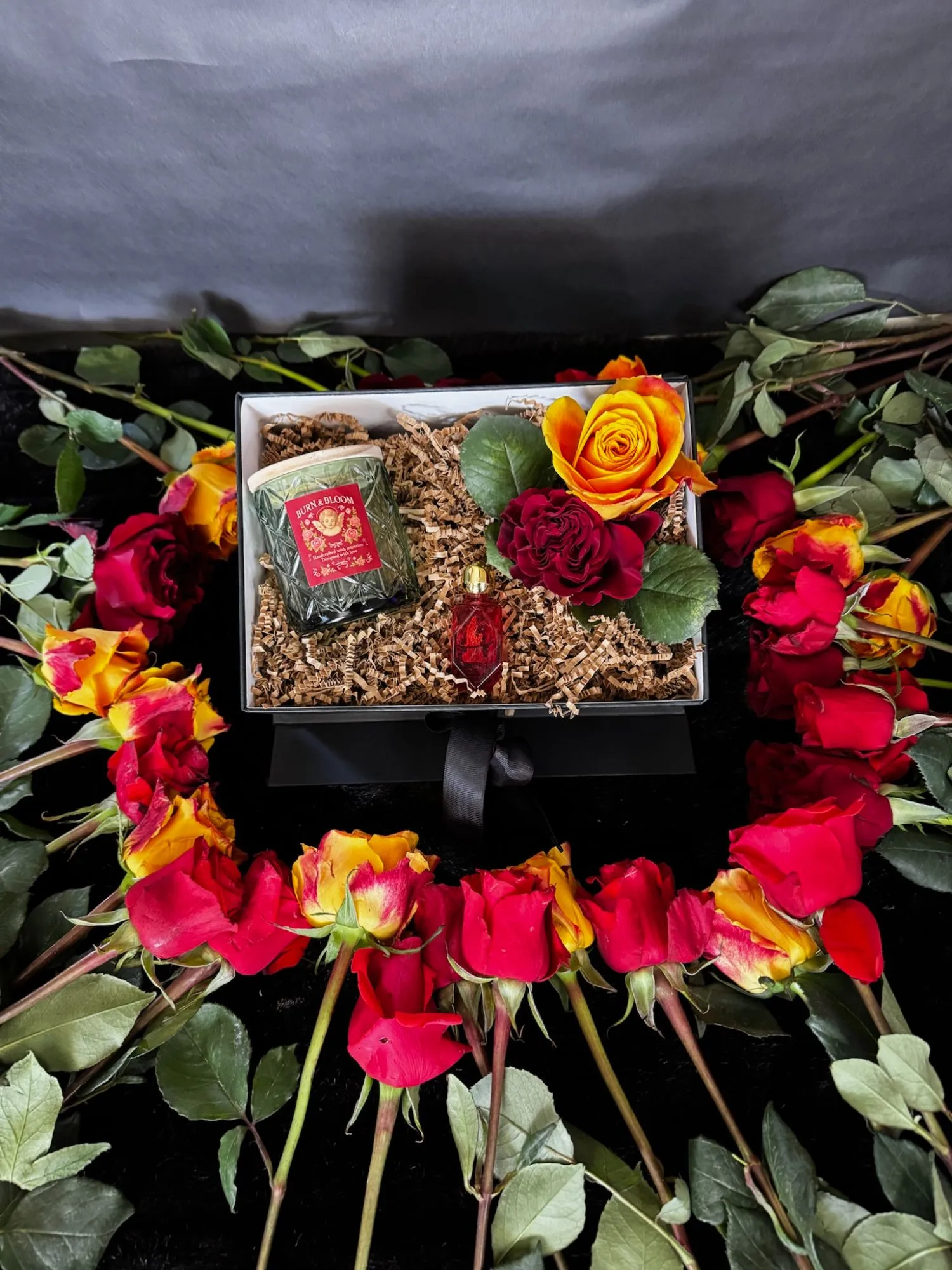 A Burn & Bloom candle and a small red perfume vial in a box among roses